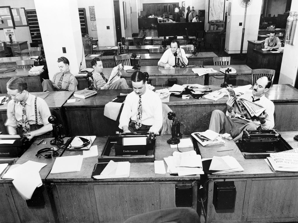 This black and white image shows the office of the New York Times circa 1942. It shows several men working at their typewriters at long wooden desks.
